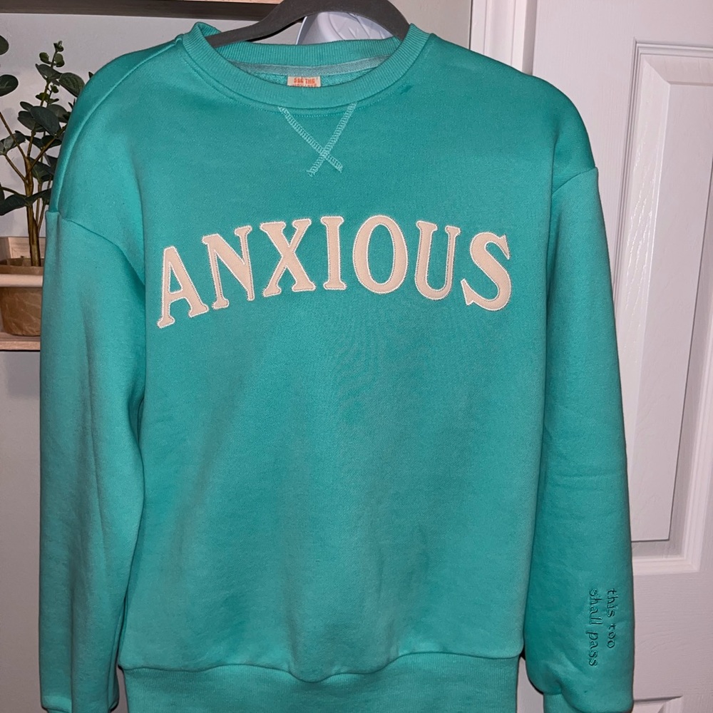 “ANXIOUS ” sweatshirt with “THIS TOO SHALL PASS” embroidered on the sleeve.